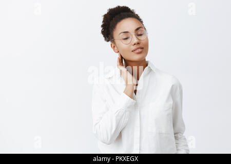 Girl rappelle baiser passionné d'amant. Portrait de l'offre africaine-américaine féminine dans les verres et chemise blanche, fermer les yeux et toucher doucement le cou, debout sur fond gris Banque D'Images