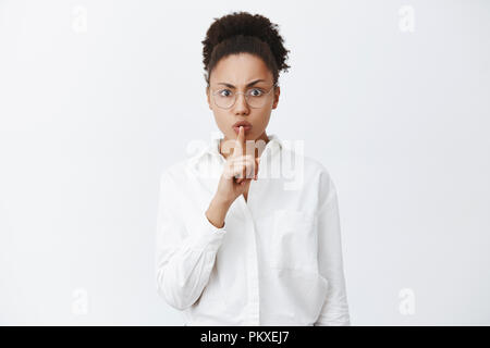 Chut, Calme, silence en classe. Le strict et le sérieux à la peau sombre enseignante en chemise col blanc et verres faisant chut geste avec l'index sur la bouche, garder la voix jusqu'exigeantes Banque D'Images