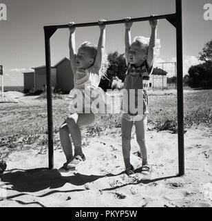 Années 1960, historiques, et deux jeunes filles summertim Holding onto et se balancer, à partir d'une barre de métal, dans une aire de jeux sur sable, England, UK. Banque D'Images