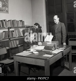 Années 1950, historique, un homme bien habillé dans une bibliothèque officiellement permanent par un tableau montrant l'intérieur d'un livre à une dame greffier assis à un bureau, England, UK. Banque D'Images