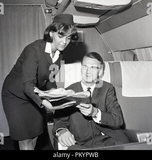 Années 1960, historiques, une femme en uniforme présentant un préposé de vol quotidien pour un homme bien passager, assis dans un siège en classe affaires sur un avion. Banque D'Images
