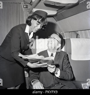 Années 1960, historiques, une femme en uniforme de bord présentant un quotidien à un passager assis dans un siège en classe affaires sur l'aéronef. Banque D'Images