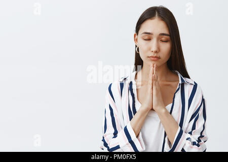 Bon à la femme entrepreneur élégant avec mignon mole sous la lèvre, fermant les yeux, détendu et permanent sur fond gris sombre dans un chemisier, tenant les mains de prier tout en rendant le souhaitent ou l'espoir Banque D'Images