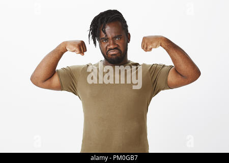 Portrait of serious-masculin à la peau sombre dodu guy dans des vêtements militaires, élever les mains et montrant les biceps, fronçant, grimaces avec expression menaçante, debout au-dessus de mur gris Banque D'Images