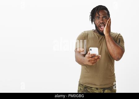 Portrait de choqué surpris à peau sombre attrayant avec guy beard dans les t-shirt et pantalon, laissant tomber la mâchoire, holding smartphone, d'être étonnés par message reçu, regardant stupéfait at camera Banque D'Images