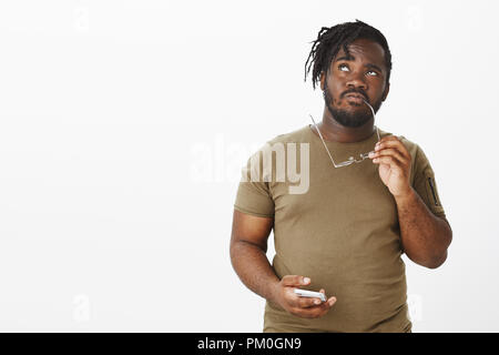 Piscine shot of African man porté en tenue décontractée, jusqu'à réfléchir ou à mémoriser, tandis que rim piqueurs de verres et holding smartphone, rappelant l'information ou la prise de décision Banque D'Images