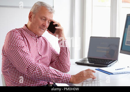 Shot de senior businessman en utilisant son téléphone portable et de faire appel alors que travailler sur ordinateur dans le bureau. Banque D'Images