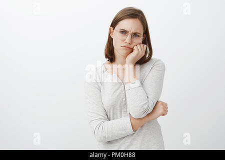 Studio shot of lonely mécontent et malheureux jeune femme dans les verres avec les cheveux bruns courts froncer et bouder de tristesse face penchée sur palm le regard avec indifférence sur mur gris Banque D'Images