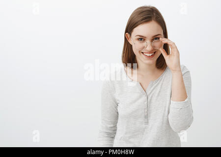 Jusqu'à la taille de balle et créative joyeuse amie intelligente avec les cheveux bruns courts à la recherche de dessous front mystérieux et intrigué, décoller les verres et un large sourire s'amuser sur mur gris Banque D'Images