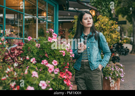 Young Pretty woman holding her smartphone et debout devant une jolie maison avec des fleurs autour. Banque D'Images