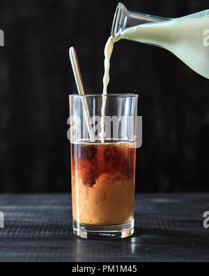 Café avec du lait. Sur une table en bois noir, le lait est versé dans un verre de café. Banque D'Images