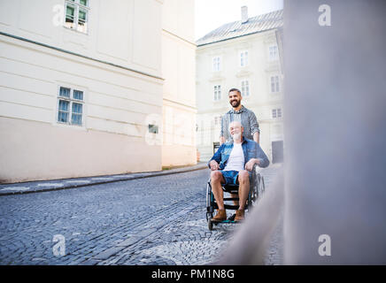 Un fils adulte avec des hauts père en fauteuil roulant, lors d'une promenade en ville. Banque D'Images
