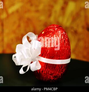 Vue de face d'un gros œuf en chocolat emballés dans du papier aluminium et rouge attaché à un noeud blanc pour Pâques sur fond jaune et noir. Selective focus Banque D'Images