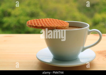 Fermé Stroopwafel placé sur le dessus de la tasse de café chaud servi sur une table en bois, feuillage vert flou en arrière-plan Banque D'Images