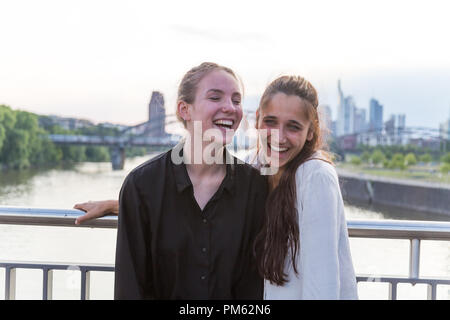 Deux jeunes femmes riant sur city bridge over river avec metropolis skyline en arrière-plan. Plan moyen. Banque D'Images