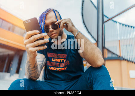 Jeune homme séduisant avec des dreadlocks bleu à la recherche à l'écran du téléphone mobile. L'homme est sur le premier plan et l'orientation, la construction en briques rouges et d'objets d'art sont sur b Banque D'Images