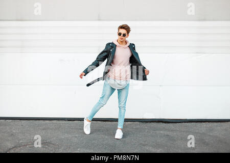 Beau jeune homme élégant avec des lunettes de soleil dans une veste en cuir noire, un t-shirt rose, bleu jeans de marque et chaussures blanches en mouvement Banque D'Images