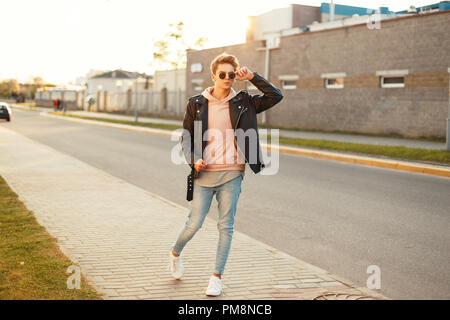 Jeune homme élégant avec des lunettes de soleil dans une veste en cuir vintage sneakers blanc et au coucher du soleil Banque D'Images