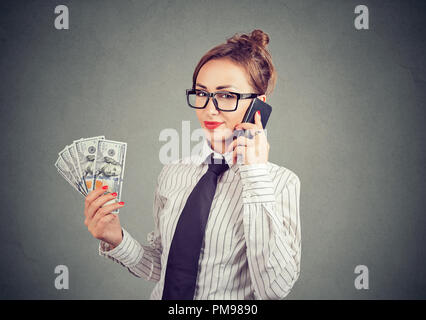 Jeune femme dans les verres satisfait holding dollar bills et parlant sur smartphone looking at camera sur fond gris Banque D'Images