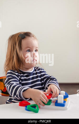 Une fille de 1,5 ans aux cheveux longs dans une robe rayée est assis à la table et joue avec un jouet en développement Banque D'Images