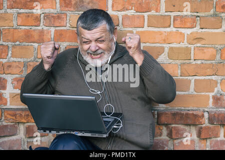 Beau portrait de senior homme barbu regardant jeu préféré sur l'écran d'un ordinateur portable alors qu'il était assis contre un mur de briques Banque D'Images