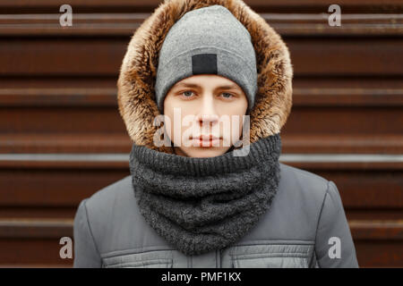 Beau jeune homme dans un manteau d'hiver à la mode avec un capuchon avec un chapeau et un foulard dans la rue Banque D'Images