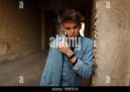 Jeune homme à la mode modèle avec une cigarette dans les vêtements en denim de fumer près d'un mur de briques sur la rue Banque D'Images