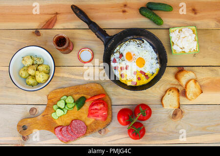 Différents aliments : oeufs brouillés dans une poêle à frire, des pommes de terre, fromage, croûtons, radis, concombres, tomates, saucisse fumée, menthe, oseille sur t en bois Banque D'Images