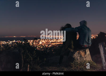 Couple en profitant de la vue d'une ville lumineuse assis au sommet d'une colline dans la nuit. Homme assis avec une femme sur une colline avec son bras autour de son épaule Banque D'Images