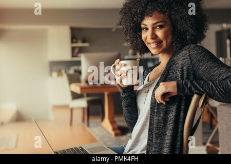 Portrait de jolie femme avec les cheveux bouclés le café à la maison avec un ordinateur portable. Femme africaine prenant pause café tout en fonctionnant de la maison. Banque D'Images