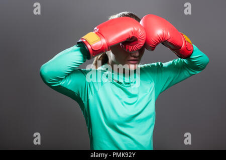 Triste jeune athlète femme aux cheveux recueillis en position debout, se tenant la main dans la boxe gants rouge près de yeux et se cacher larmes après coup. Piscine Banque D'Images