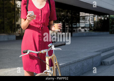 Femme en robe à pois avec transport de vélo, contrôle de courrier en ligne et le café. Personne de sexe féminin à l'aide de smartphone et holding tasse de boisson chaude sur Banque D'Images