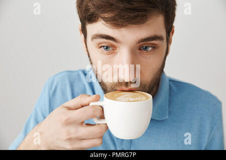 Close-up shot of cute hipster barbu en sirotant un cappuccino tout en travaillant dans un café en tant que travailleur indépendant, bénéficiant d'une atmosphère agréable et d'être concentré, sur fond gris. Femme fait de lui au matin café Banque D'Images