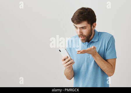 Qui vous a enseigné la grammaire. Studio shot of agacé et expressive portrait guy avec barbe holding smartphone tout en regardant l'écran et main tremblante, l'expression de la gêne et de la confusion Banque D'Images