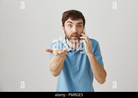 Les émotions et la technologie concept. Choqué et impressionné caucasian guy talking on cellphone tout en dirigeant à l'appareil photo avec l'expression surprise, debout sur fond gris. Recherche rapide, me donner pen Banque D'Images