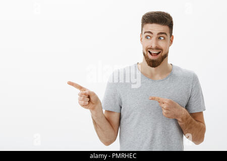 Guy enthousiaste se sentir heureux et surpris à regarder l'espace copie super cool. Portrait of handsome ravis et homme charismatique avec barbe et moustache à la recherche et pointant vers la gauche sur mur gris Banque D'Images