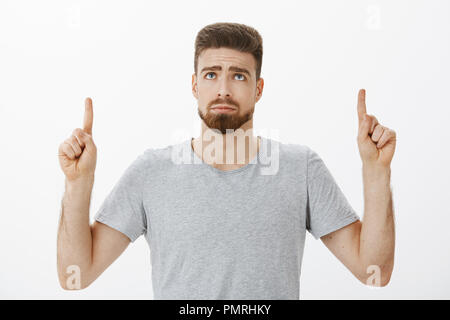 Triste et beau mignon timide adultes masculin homme avec barbe et des yeux bleus à la pointe vers le haut et de froncer regardant sombre sentiment injuste chose, rester envieux et pissé sur mur gris Banque D'Images