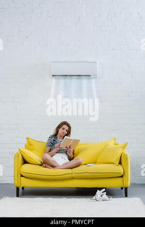 L'accent young woman reading book on couch par pendaison de climatiseur sur le mur et le souffle de l'air froid Banque D'Images