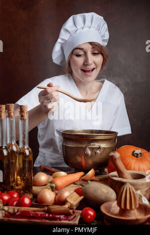 Belle jeune fille dans un uniforme de chef avec de vieux pan en laiton et cuillère en bois nourriture d'échantillon. Sur une table divers ustensiles de cuisine et les légumes. Banque D'Images
