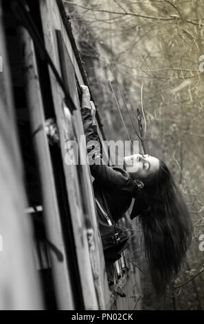 Jeune brunette woman hanging out d'une fenêtre de train abandonné dans les bois avec de longs cheveux rouges Banque D'Images