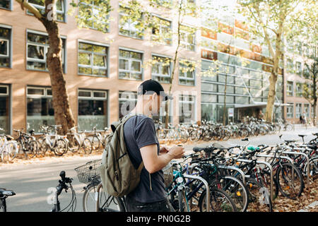 Un étudiant avec un sac à dos ou un touriste sur la rue de Leipzig en Allemagne utilise un téléphone cellulaire à côté du parking à vélos qui est à côté de la bibliothèque de l'Université de Leipig et student hostel. Banque D'Images