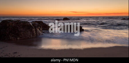 La marée de l'océan change pendant le coucher du soleil avec quelques rochers dans le sable. Banque D'Images