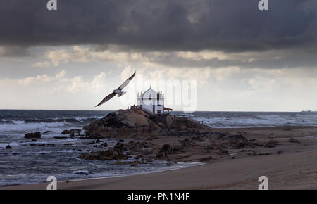 À la plage, une mouette vole avec une chapelle construite sur les rochers en arrière-plan, pendant un jour de tempête. Banque D'Images