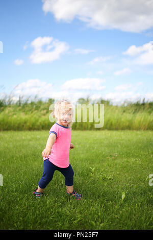 Un mignon petit enfant de deux ans est fille de rire alors qu'elle tourne autour et joue à l'extérieur dans l'herbe verte sur un jour d'été venteuse. Banque D'Images