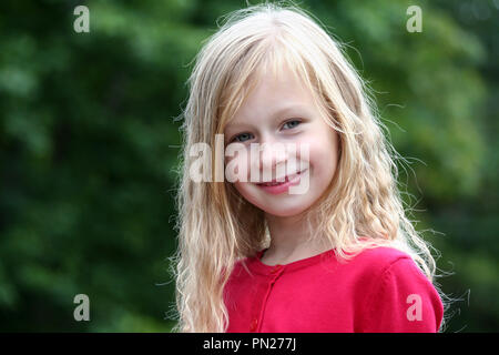 Portrait petite fille aux longs cheveux blonds dans un chandail rouge et souriant à directement à l'appareil photo, à l'arrière-plan vert des arbres, son feuillage foncé, Banque D'Images