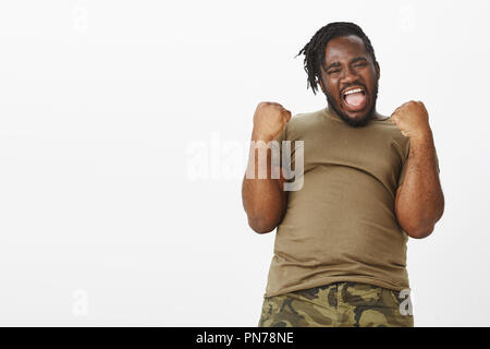 Ouais c'est ce que nous avons fait. Portrait of happy énergique de peau foncée guy dodus dans t-shirt militaire, élever les mains avec les poings serrés, se pencher en arrière et tout sourire, célébrant la victoire, étonné avec win Banque D'Images