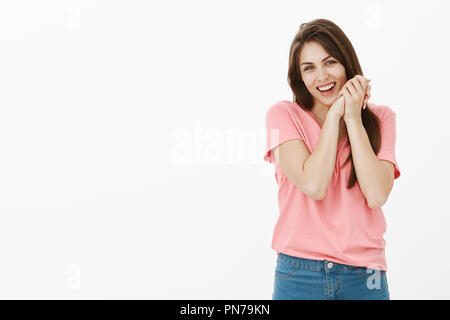 Tourné à l'intérieur de touché et heureux de l'adorable modèle féminin en t-shirt rose, tilting head holding et serra les mains près de visage tout en sourire, d'être ému par quelque chose de tendre et surprenant Banque D'Images