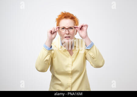 Caucasian woman attentif avec de courts cheveux rouges ressemble à distance, garde scrupuleusement part sur la jante de spectacles. Essayez de voir quelque chose de très loin, l'isolat Banque D'Images