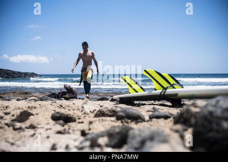 Jeune adolescent male surfer qui sort de l'eau après une session de surf dans les vagues de l'océan. table et des ailerons sur les rochers. locations et de l'eau sport ac Banque D'Images