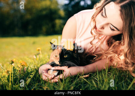 Girl lying on grass avec cat. Printemps ou été concept par temps chaud. Arrière-plan flou. Ginger kitten avec deux visage masque de couleur. Banque D'Images